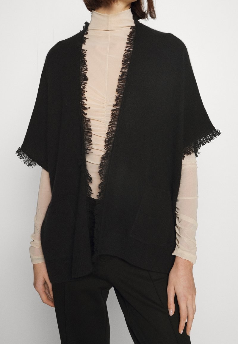 Personne portant un haut à col roulé beige transparent sous un cardigan noir à manches courtes avec des franges sur les bords et un pantalon noir, debout contre un fond blanc.