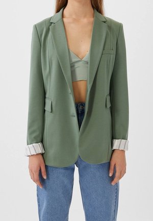 Persoon draagt een lichtgroene blazer met opgerolde gestreepte manchetten, een bijpassende groene cropped top eronder en een blauwe spijkerbroek.