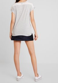 Kvinna som står, iklädd en grå kortärmad t-shirt, svart minikjol och vita sneakers, vänd bort mot en enfärgad bakgrund.