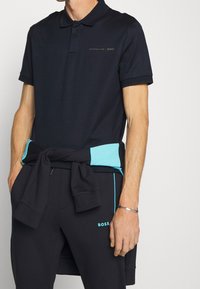 Polo shirt marine avec une finition texturée, présentant un logo subtil et des accents turquoises, associé à un pantalon de survêtement assorti et un sweat-shirt noué à la taille.