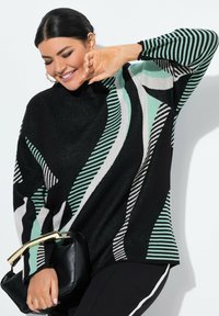 Femme souriante aux cheveux noirs portant un pull rayé noir, vert et blanc, tenant un sac à main noir avec une anse en or sur fond blanc.