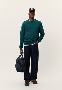 Φούτερ σε χρώμα τιλ, σκουρόχρωμο τζιν, καφέ παπούτσια, κρατώντας μια καρό τσάντα. Όλα τα ρούχα έχουν χαλαρή εφαρμογή με διακριτικές λεπτομέρειες και υφές.