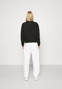 Svart sweatshirt med rund hals och ribbade ärmar, tillsammans med vita löst sittande byxor och vita sneakers. Bakgrunden är enkel.