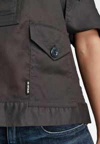 Chemise gris foncé à manches courtes avec poche à boutons et étiquette en tissu noire G-STAR, portée avec un jean en denim bleu.