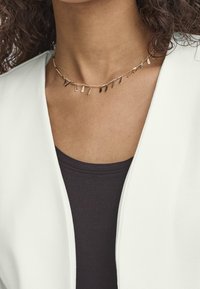 Gros plan d'une personne portant un collier chaîne en or avec petits charms rectangulaires pendants, associé à un blazer blanc et un haut noir.