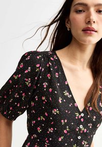 Robe noire à manches courtes bouffantes, avec des motifs floraux roses et des boutons blancs. Fabriquée en tissu léger avec un décolleté en V profond.