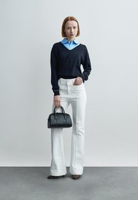 Pull noir à col en V sur une chemise bleu clair, jeans blancs évasés, chaussures à talons marron et un petit sac à main noir.