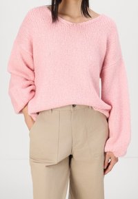 Maglione rosa oversize a maglia con scollatura ampia e tessuto texture, abbinato a pantaloni beige a vita alta con tasche laterali.
