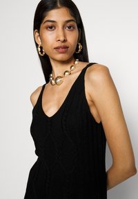 Black knitted tank top with a V-neck and diamond pattern. Accessories include large gold hoop earrings and a chunky gold necklace.