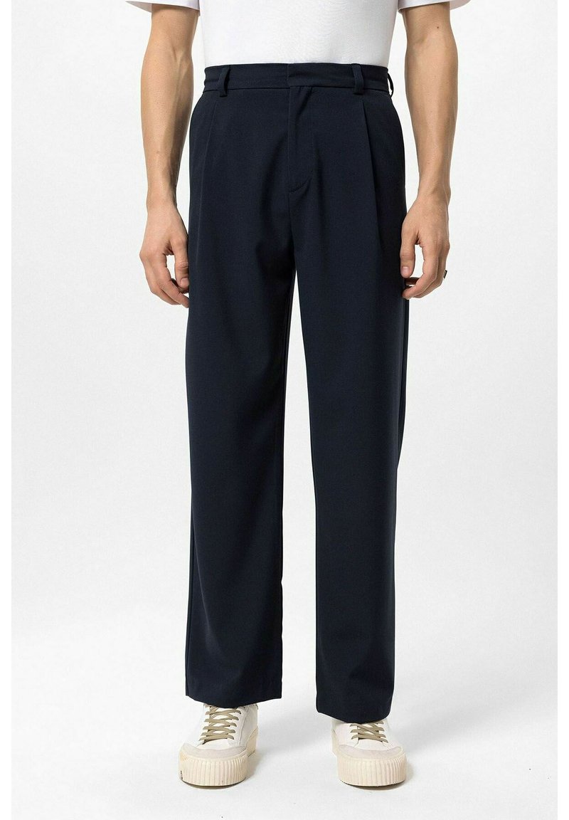 Marineblauwe plisségordelbroek gemaakt van een gladde stof, met een getailleerde pasvorm, zijzakken, en gedragen met lichtgekleurde sneakers.