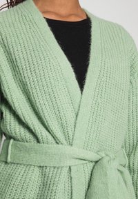 Miętowozielony sweter z dzianiny o strukturalnym wzorze, z otwartym przodem i paskiem do związania. Nałożony na czarny top w prążki.