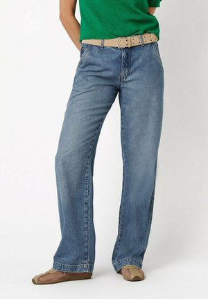 Person trägt hellblaue Jeans mit weitem Bein, beigen Gürtel, grünes Oberteil und beige Mesh-Schuhe, steht vor einem einfarbigen weißen Hintergrund.