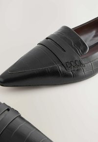 Schwarze Lederloafer mit einem strukturierten Krokodilmuster, spitzer Zehenform und einem dekorativen Riemen an der Vorderseite mit gestickten Akzenten.