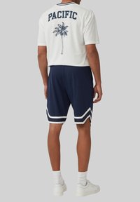 Witte t-shirt met korte mouwen met "PACIFIC" en palmboomafbeelding op de achterkant, gecombineerd met marineblauwe shorts met witte zijstreep. Witte sneakers.