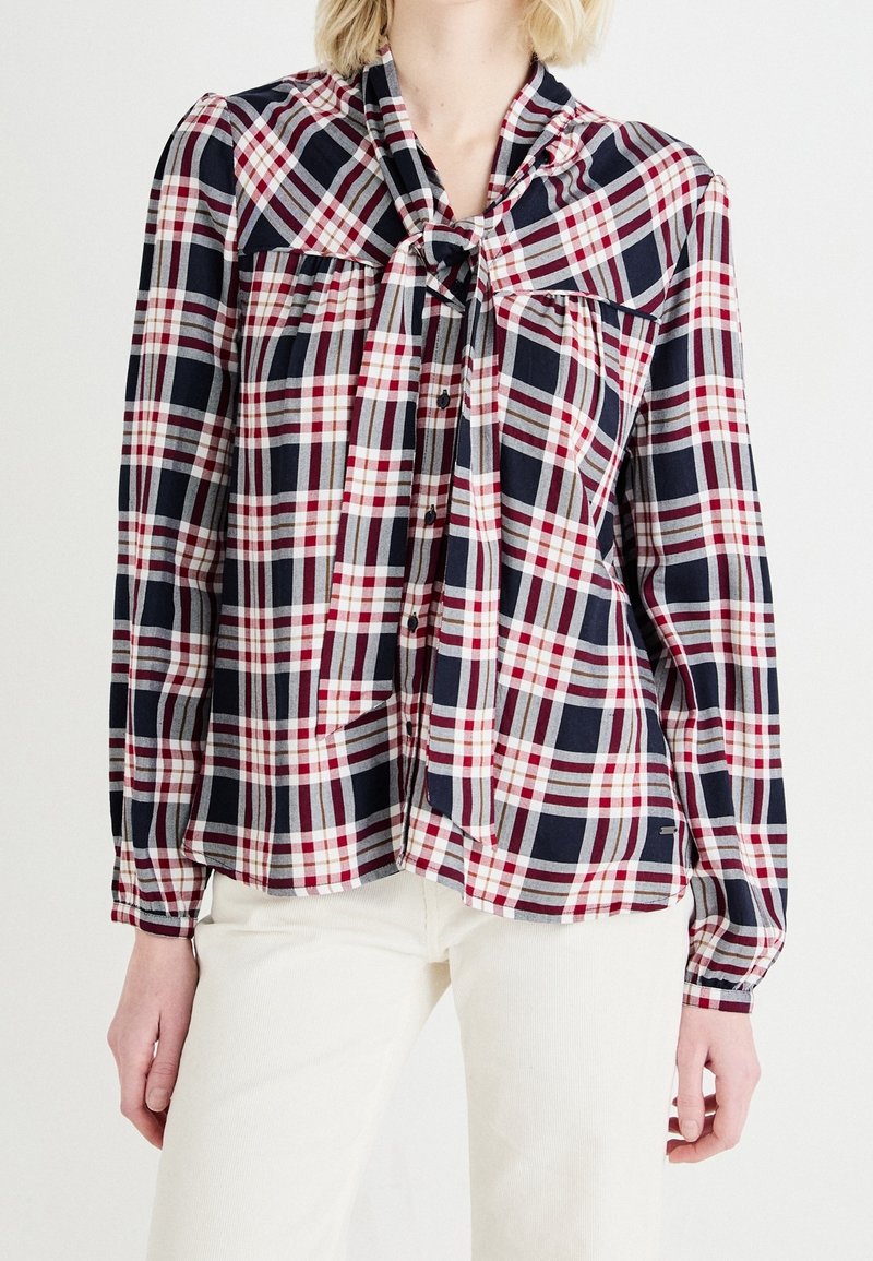 Femme portant un chemisier à manches longues à carreaux bleu marine, blanc et rouge avec un nœud à l'avant, associé à un pantalon écru.