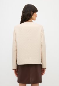 Ljusbeige quiltad jacka med ett geometriskt mönster, rund halsringning, långa ärmar och brun kant vid ärmsluten, tillsammans med en brun läderskjorta.