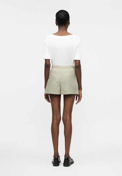 Γυναίκα με σκούρο δέρμα και μαλλιά σε κότσο, φοράει λευκή μπλούζα με κοντά μανίκια, ανοιχτό γκρι σορτς με ρίγες και μαύρα μοκασίνια, κοιτάζοντας προς τα πίσω σε λευκό φόντο.