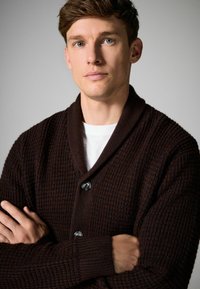 Mörkbrun texturerad cardigan med sjalkrage, med två knappar och ribbade ärmavslut, som bärs över en vit skjorta, med korsade armar.