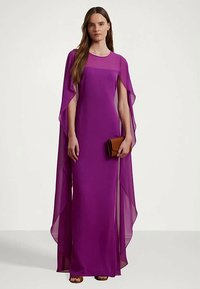 Sieviete, ģērbusies garā purpura kleitā ar plūstošām apmetņu piedurknēm, tur brūnu rokassomu un stāv pretī vienkāršam fonam.