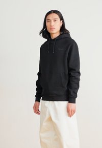Felpa nera con cappuccio, tasca frontale, polsini e orlo a coste, e cordini; indossata con pantaloni bianchi larghi.
