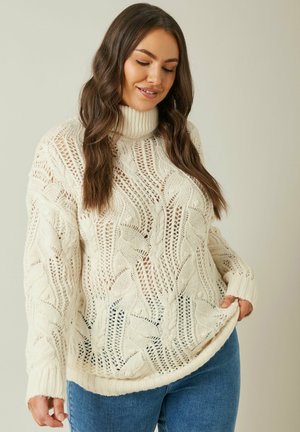 Maglione crema a trecce con collo alto, caratterizzato da intricati motivi a giorno e polsini e orlo a coste. Indossato sopra un paio di jeans blu.