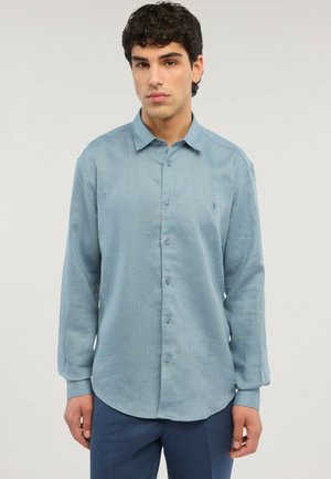 Junger Mann, der ein hellblaues Hemd mit langen Ärmeln und dunkle blaue Hose trägt, vor einem schlichten hellen Hintergrund steht.