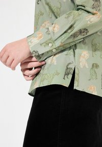 Camisa verde claro de tela con un estampado de animales en negro y naranja. Mangas largas con cierres de botón. Rendis de lado visibles.