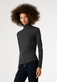 Görbe hajú nő fekete garbós felsőben, fehér pöttyökkel és sötétkék farmernadrágban, egy sima világos háttér előtt áll.