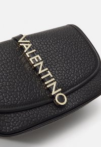 Černá texturovaná kožená kabelka s zakřiveným tvarem, s výrazným zlatým logem "VALENTINO" na přední straně.