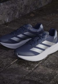 Modré športové topánky s priedušným sieťovinovým zvrškom so striebornými akcentmi a tromi pruhmi, s bielou polstrovanou medzipodrážkou a textúrovanou podrážkou.