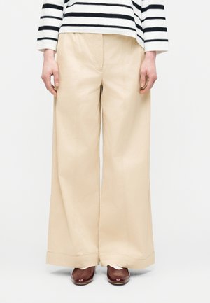 Osoba oblečená v béžových nohaviciach s širokými nohavicami, bielom tričku s dlhým rukávom a čiernymi pruhmi a hnedých topánkach s okrúhlym nosom.