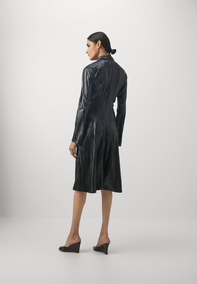 Robe noire à manches longues en tissu texturé et brillant. Elle présente un corsage ajusté, une jupe évasée et un col. Portée avec des mules à talons marron.