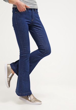 Person iført mørkeblå vidde jeans, grå langærmet top og metallic slip-on sneakers mod en ensfarvet hvid baggrund.