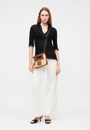 Žena oblečená v černém průsvitném topu, bílých kalhotách s širokými nohavicemi, černých botách na podpatku s otevřenou špičkou a nesoucí malou hnědo-béžovou kabelku přes rameno.