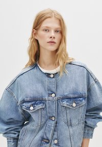 Svetlo modra jeans jakna z gumbnim zapiranjem, dvema žepoma na prsih, oversized krojem in razfrakanimi detajli ovratnika. Gladka tekstura.