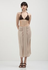 Femme portant un haut de bikini marron, une jupe enroulée beige, des sandales marron et des boucles d'oreilles en or, se tenant devant un fond clair.