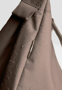 Borsa in tessuto marrone resistente all'acqua con gocce visibili, caratterizzata da una superficie texture e un accento metallico vicino alla cucitura.