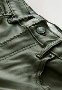 Denim verde oliva con una textura suave, que presenta un bolsillo frontal, un botón de metal oscuro y costuras contrastantes a lo largo de las costuras.