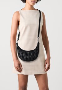 Sac à bandoulière en cuir synthétique noir avec des plis diagonaux texturés, quincaillerie argentée et une bandoulière réglable, porté avec une robe à carreaux beiges.
