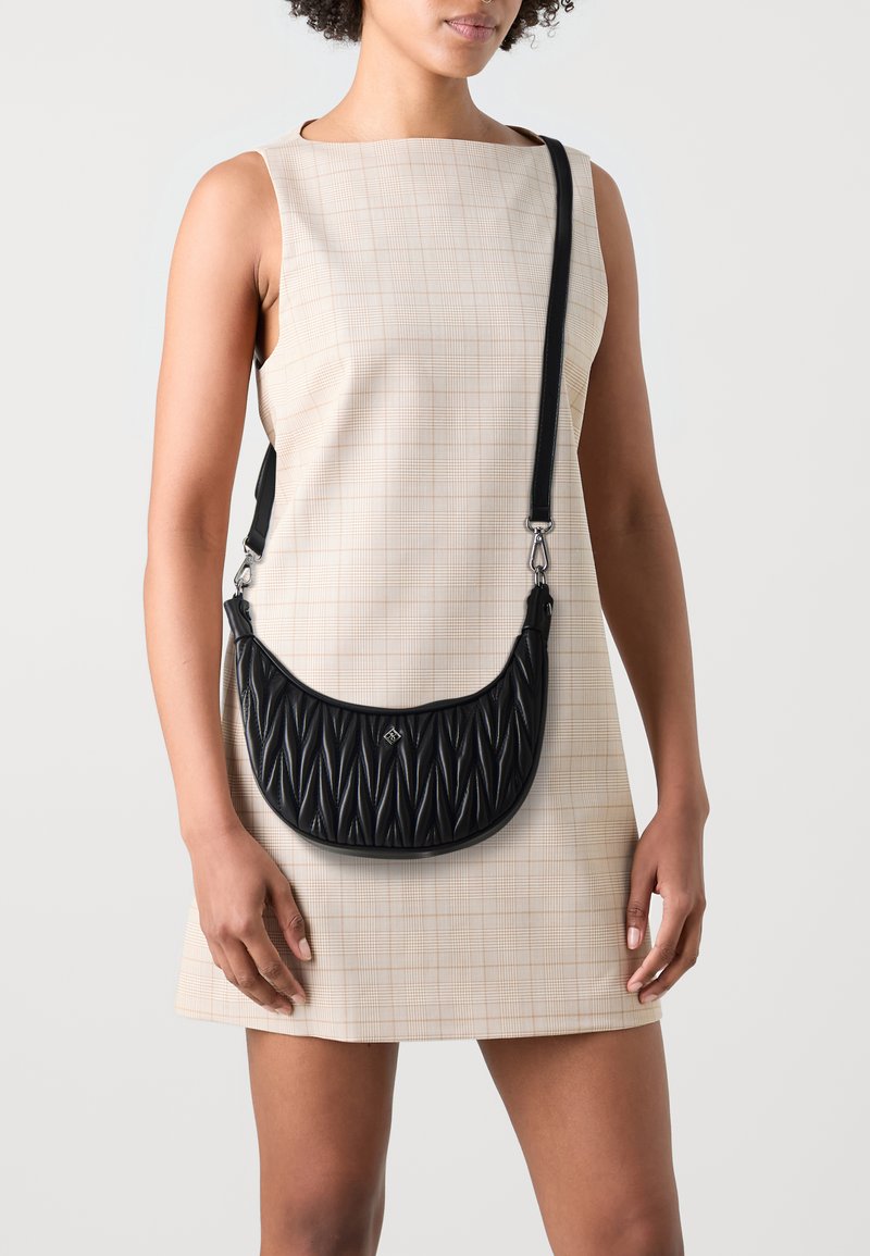 Sac à bandoulière en cuir synthétique noir avec des plis diagonaux texturés, quincaillerie argentée et une bandoulière réglable, porté avec une robe à carreaux beiges.