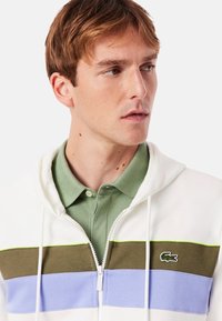 Sudadera con capucha en blanco, con rayas horizontales en verde, oliva y azul. Cremallera frontal y logotipo de Lacoste en el pecho. Material de mezcla de algodón.