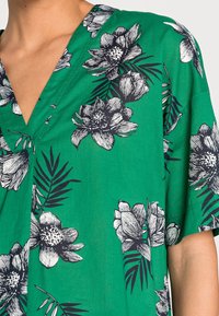 Prenda verde con escote en V, que presenta patrones de flores y hojas en negro. La tela parece ligera y tiene una textura suave.