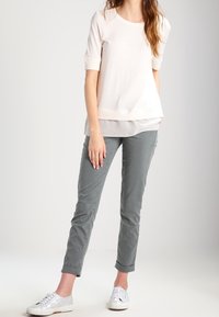 Jasnoróżowa bluzka z krótkim rękawem o warstwowym designie, noszona z dopasowanymi szarymi spodniami i białymi sneakersami. Gładkie tekstury, luźny krój.