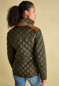 Maslinasto zeleni jakna od kvadratića s visokim ovratnikom, koja ima smeđe antilopske naglaske na ramenima i podesiv struk s kopčama.