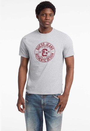 Gray cotton t-shirt featuring a red circular logo with "GUESS JEANS," "AUTHENTIC DESIGN," and an eagle graphic. Short sleeves, classic fit.