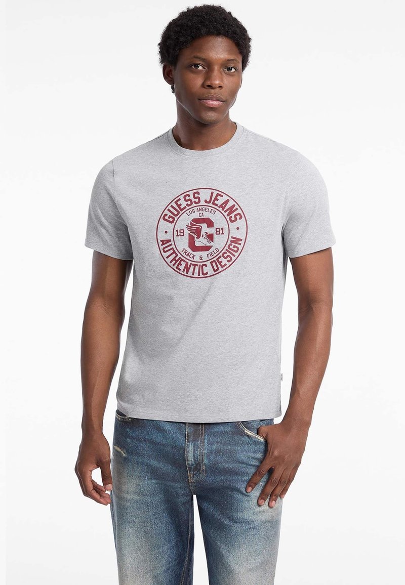 Grå bomulds t-shirt med et rødt cirkulært logo, der har "GUESS JEANS," "AUTHENTIC DESIGN," og en ørne-grafik. Korte ærmer, klassisk pasform.