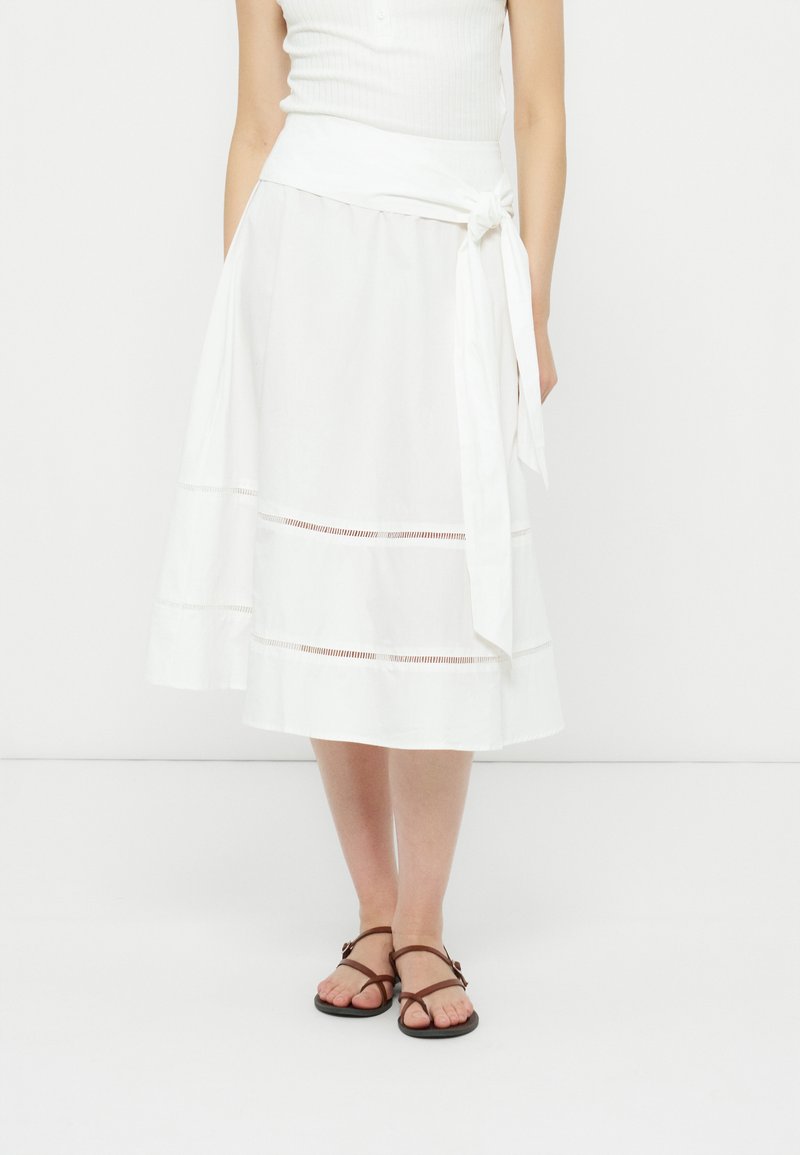 Femme portant une jupe portefeuille midi blanche avec des détails découpés horizontaux et des sandales plates marron, debout devant un fond clair uni.