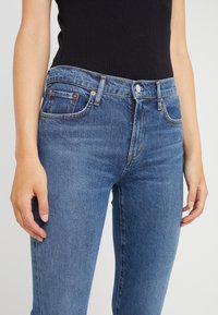 Mörkblå denimjeans med en figursydd design, fem fickor, kopparnitar och en klassisk knappstängning. Slät textur med subtila blekningar.