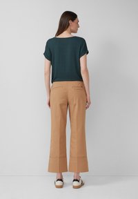 Donker teal kortek fonte top gecombineerd met zandkleurige wijd-uitlopende broeken met twee achterzakken en een cropped zoom. Draagt beige sneakers.