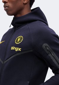 Άνδρας που φοράει σκούρο μπλε μπουφάν Chelsea FC με φερμουάρ, με κίτρινο λογότυπο συλλόγου, όνομα χορηγού BingX και κλειδωμένη τσέπη στο αριστερό μανίκι.